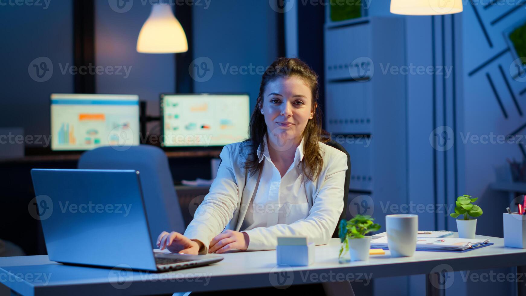 Business woman looking at camera smiling after typing on laptop sitting at desk in start-up company late at night. Focused employee using technology network wireless doing overtime for job reading. photo