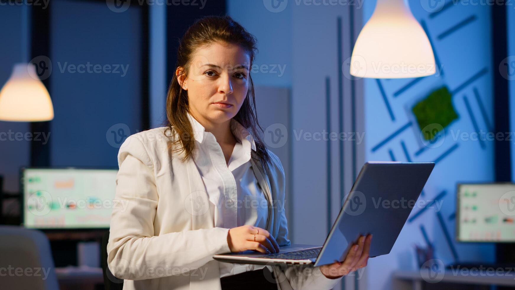 Manager woman holding laptop, typing and looking satisfied at camera standing in start-up company late at night. Focused employee using technology network wireless doing overtime for job reading. photo