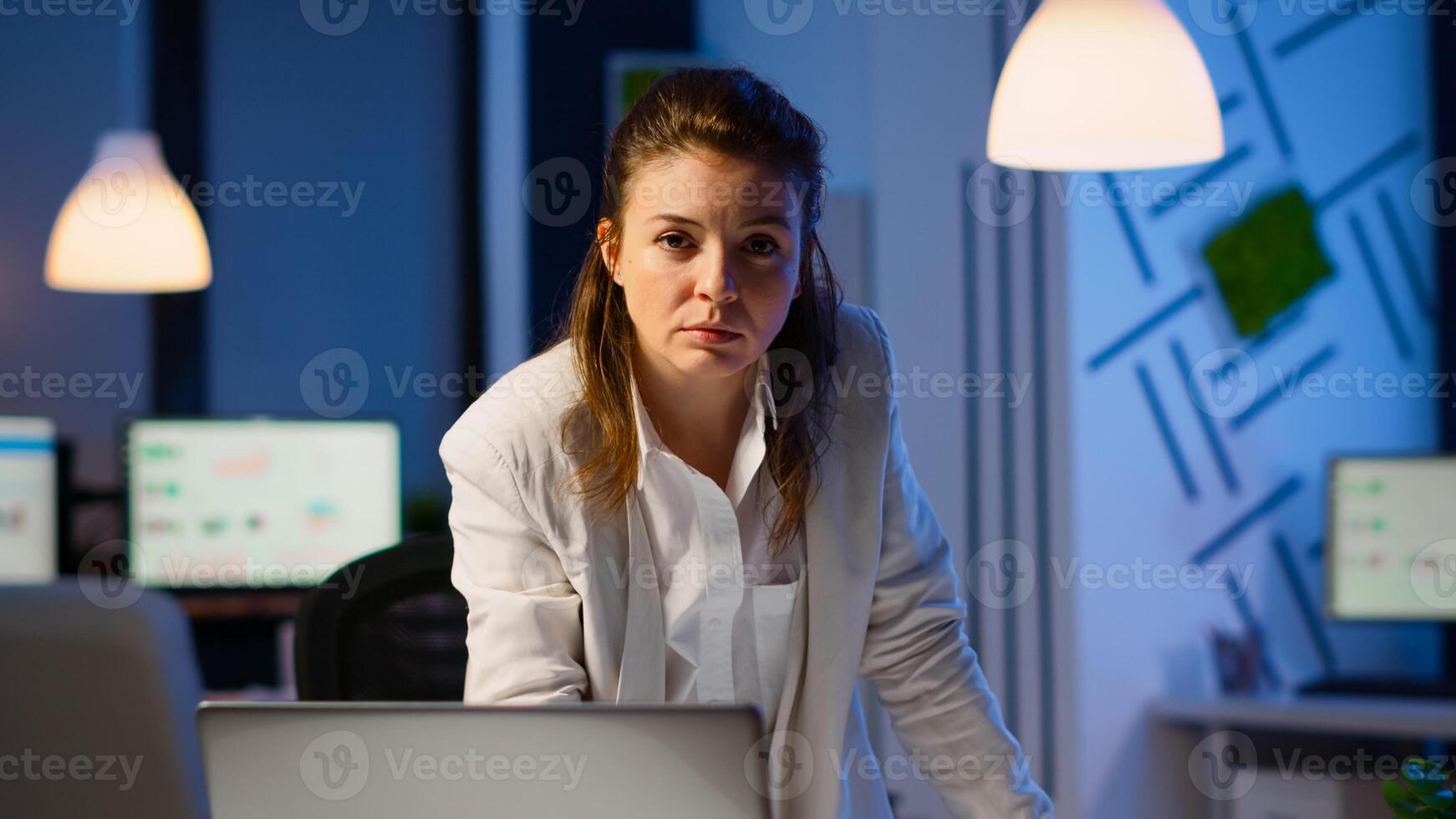 Focused tired business woman looking at camera after reading mails on laptop standing near desk in start-up company late at night. Focused employee using technology network wireless doing overtime photo