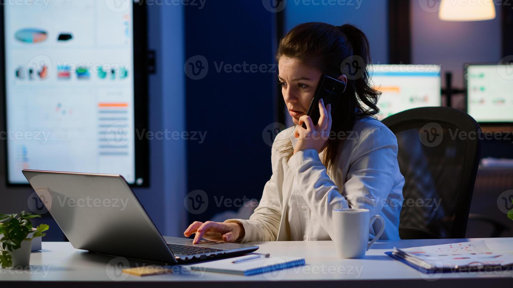 Woman employee speaking at phone while working at laptop late at night. Busy focused freelancer using modern technology network wireless doing overtime for job reading writing, searching taking break photo