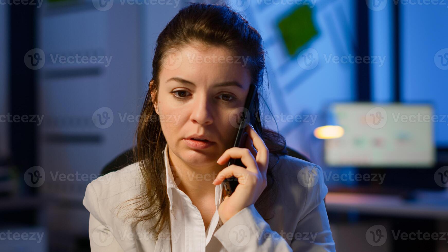 Concentrated businesswoman during phone call late at night working from business office discussing with coworker. Busy employee using modern technology network wireless doing overtime for job talking photo