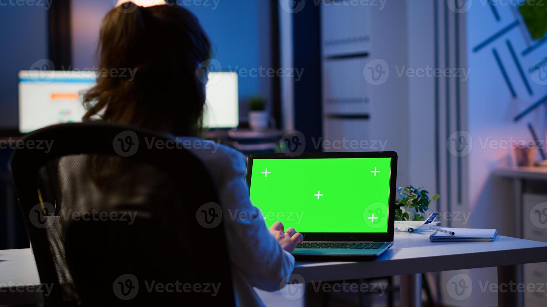 Back view of business woman looking at green screen laptop sitting at desk in start up office overworking. Freelancer watching desktop monitor display with green mockup, chroma key, during night time photo