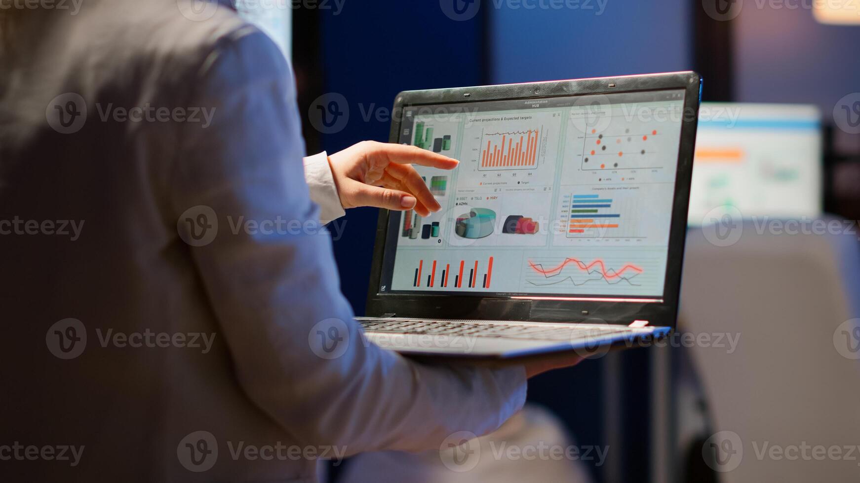 Back view of business woman analysing financial statistics holding laptop standing in start-up office late at night overworking. Busy focused employee using modern technology network wireless overtime photo