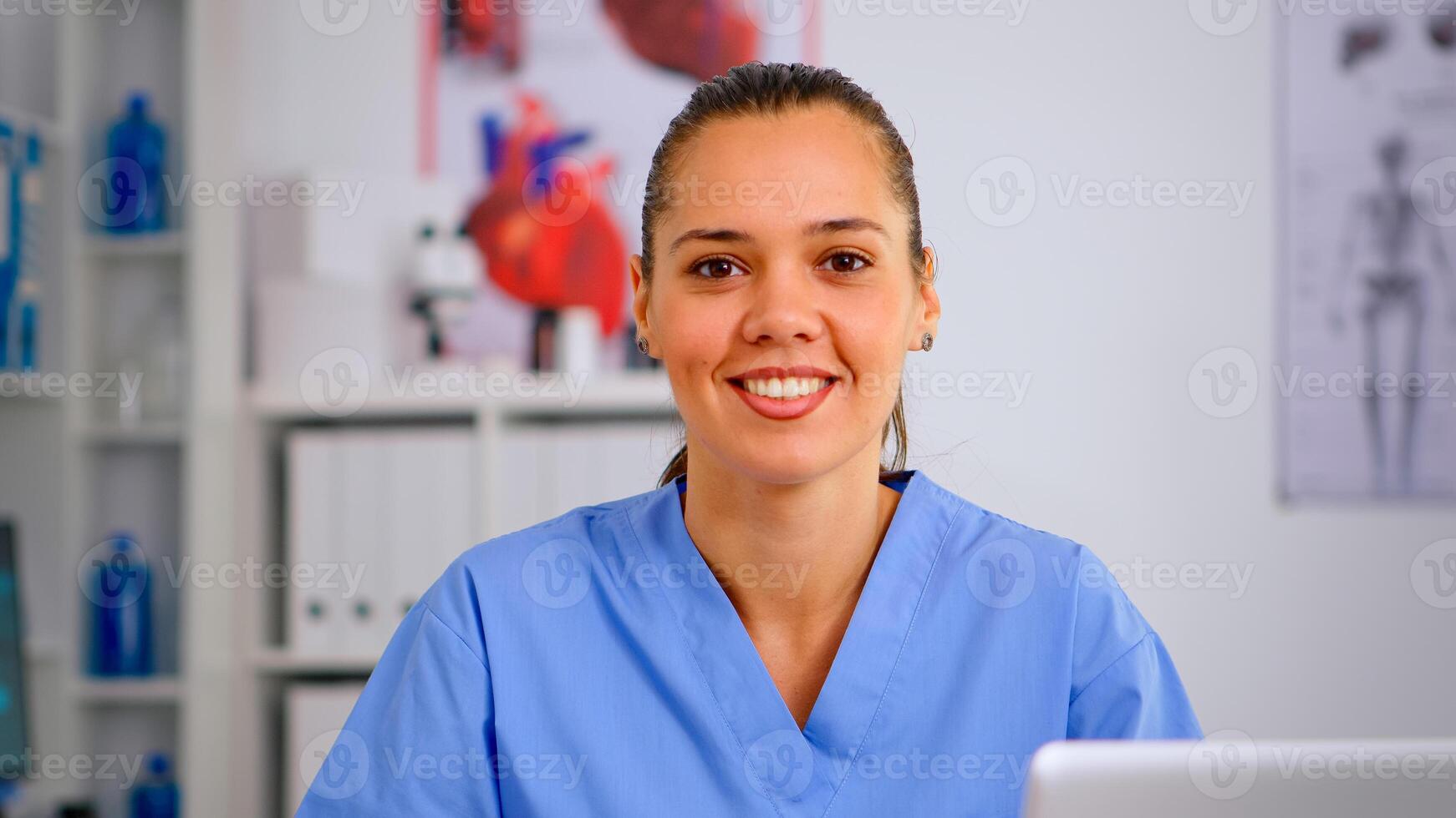 Portrait of nurse looking at camera smiling after typing on laptop sitting in hospital office wearing blue uniform. Hospital healthcare worker, medicine clinical assistant health consultant therapist photo