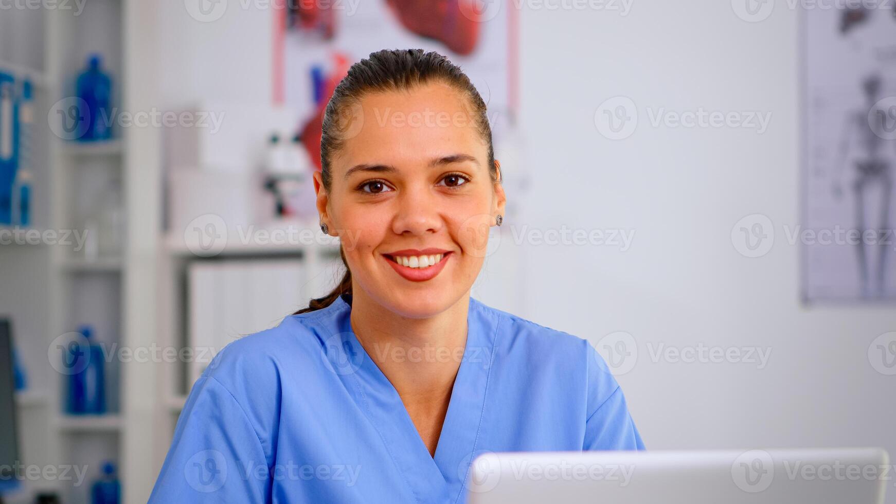 Close up of young medical assistant typing at laptop and raising head smiling at camera sitting in hospital office wearing blue uniform. Hospital healthcare worker, medicine clinical assistant health consultant therapist photo