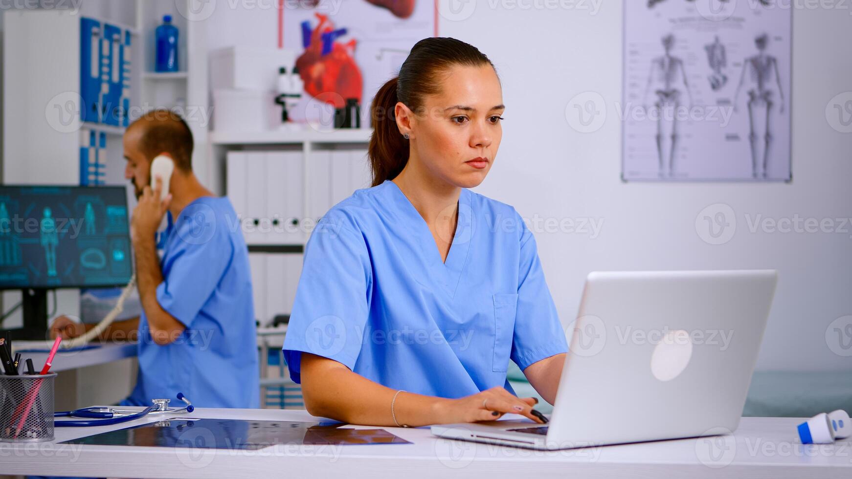 Medical practitioner typing patient health report on laptop in hospital office. Health care staff sitting at desk talking at phone in background, using computer in clinic looking at monitor, medicine. photo