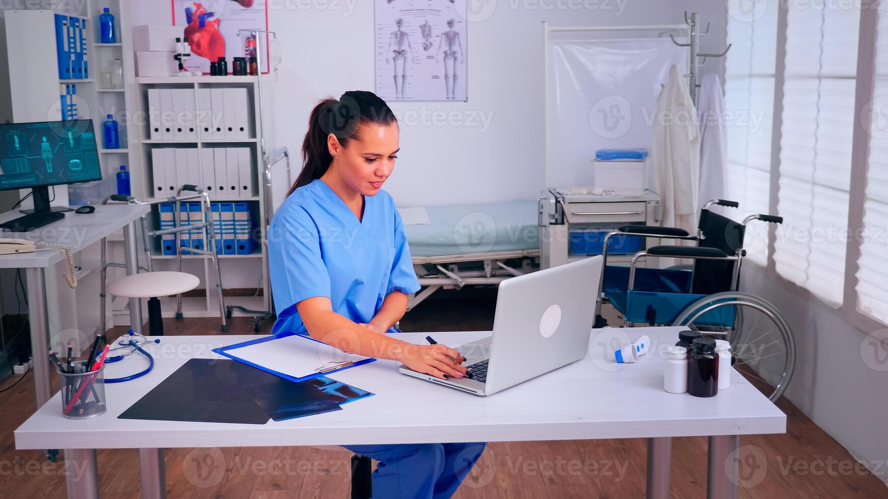 Therapist writing list of consulted patients online, making research, noting, taking useful information on clipboard. Doctor assistant in uniform working on laptop in modern hospital office. photo