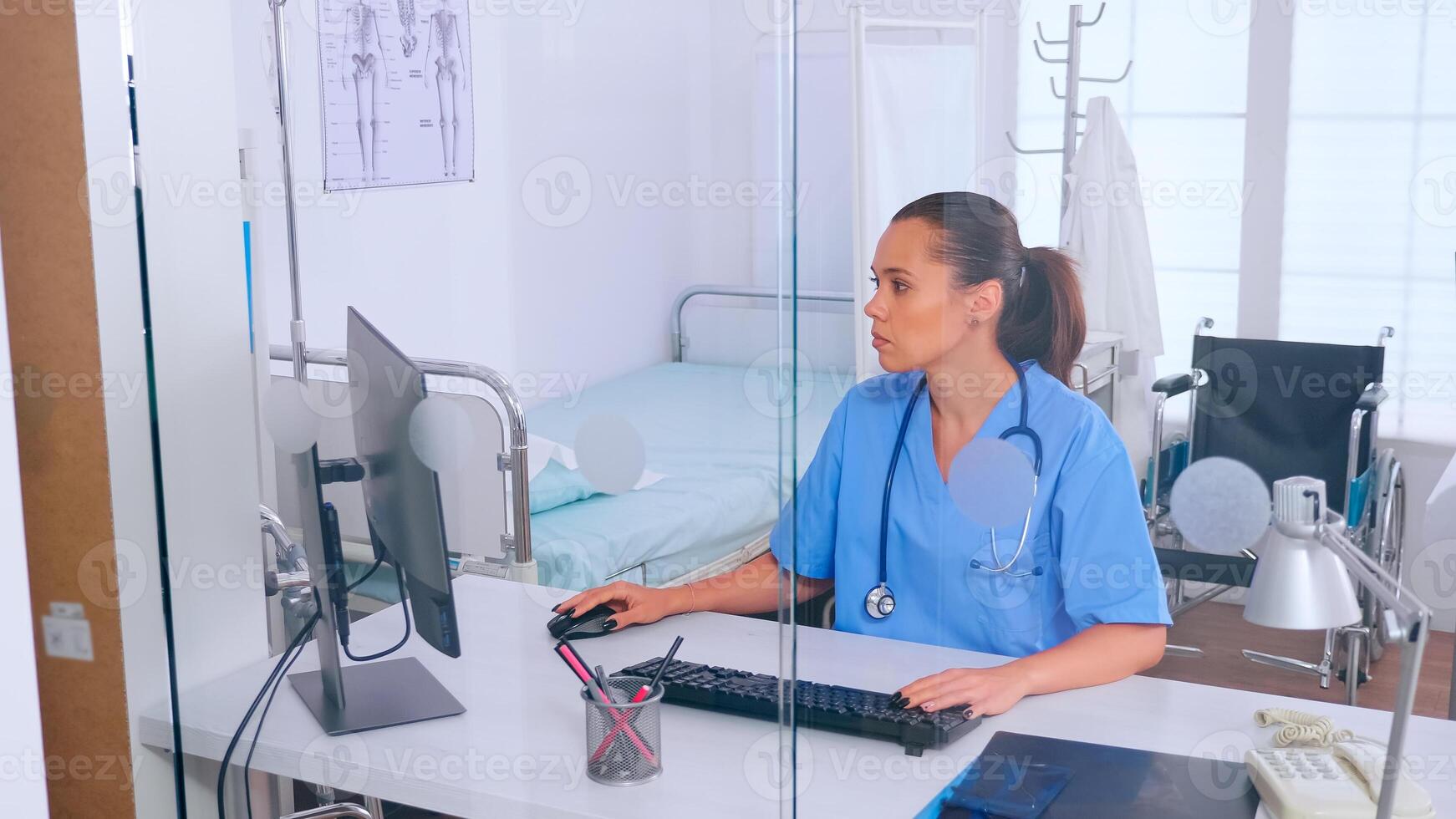 Doctor assistant typing on computer with a monitor sitting behind a glass wall in the office. Medical physician in medicine uniform writing list of consulted, diagnosed patients, making research. photo