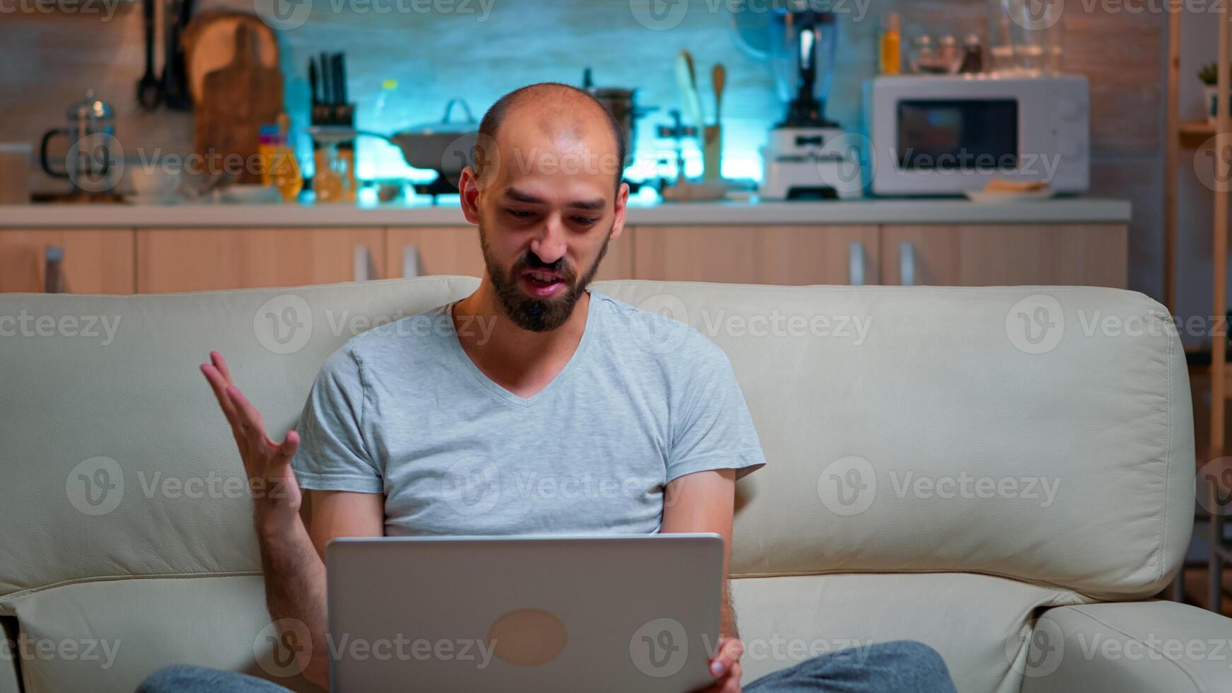 Caucasian male sitting on couch in pajamas working on laptop computer during videocall conference. Comfortable man relaxing on sofa chatting with business people late at night in kitchen photo