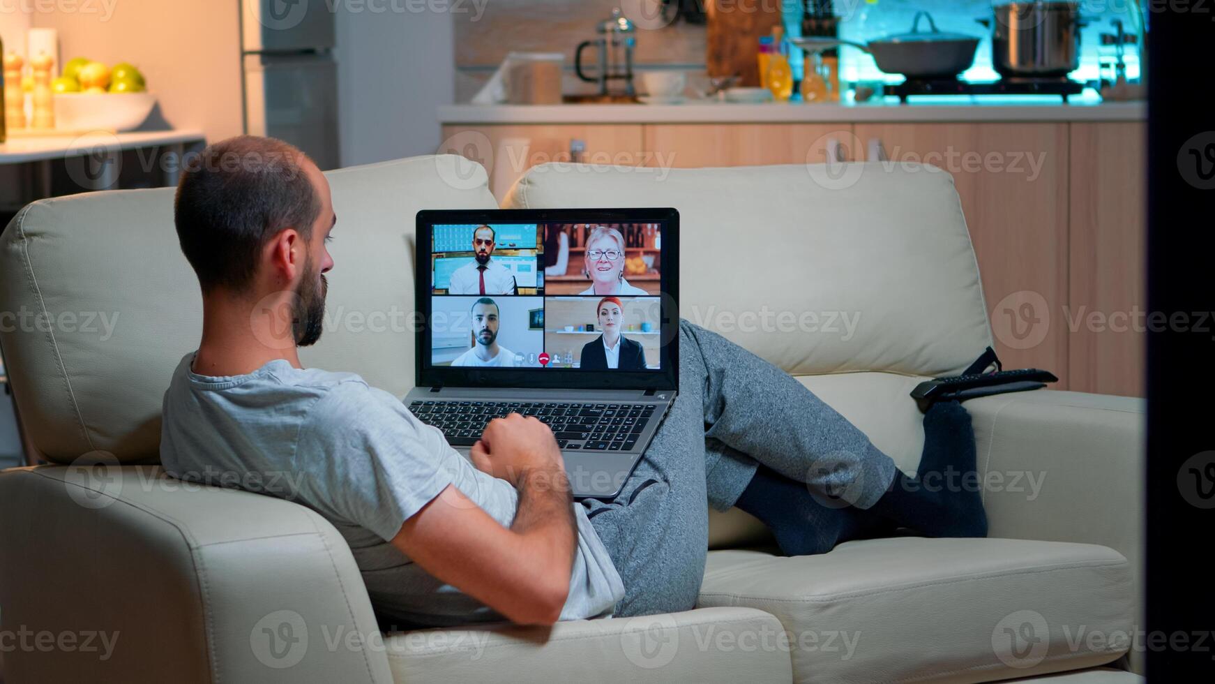 Comfortable man relaxing on couch while having online business conference with collegues using laptop computer during quarantine. Focused adult talking on videocall use webcam late at night in kitchen photo