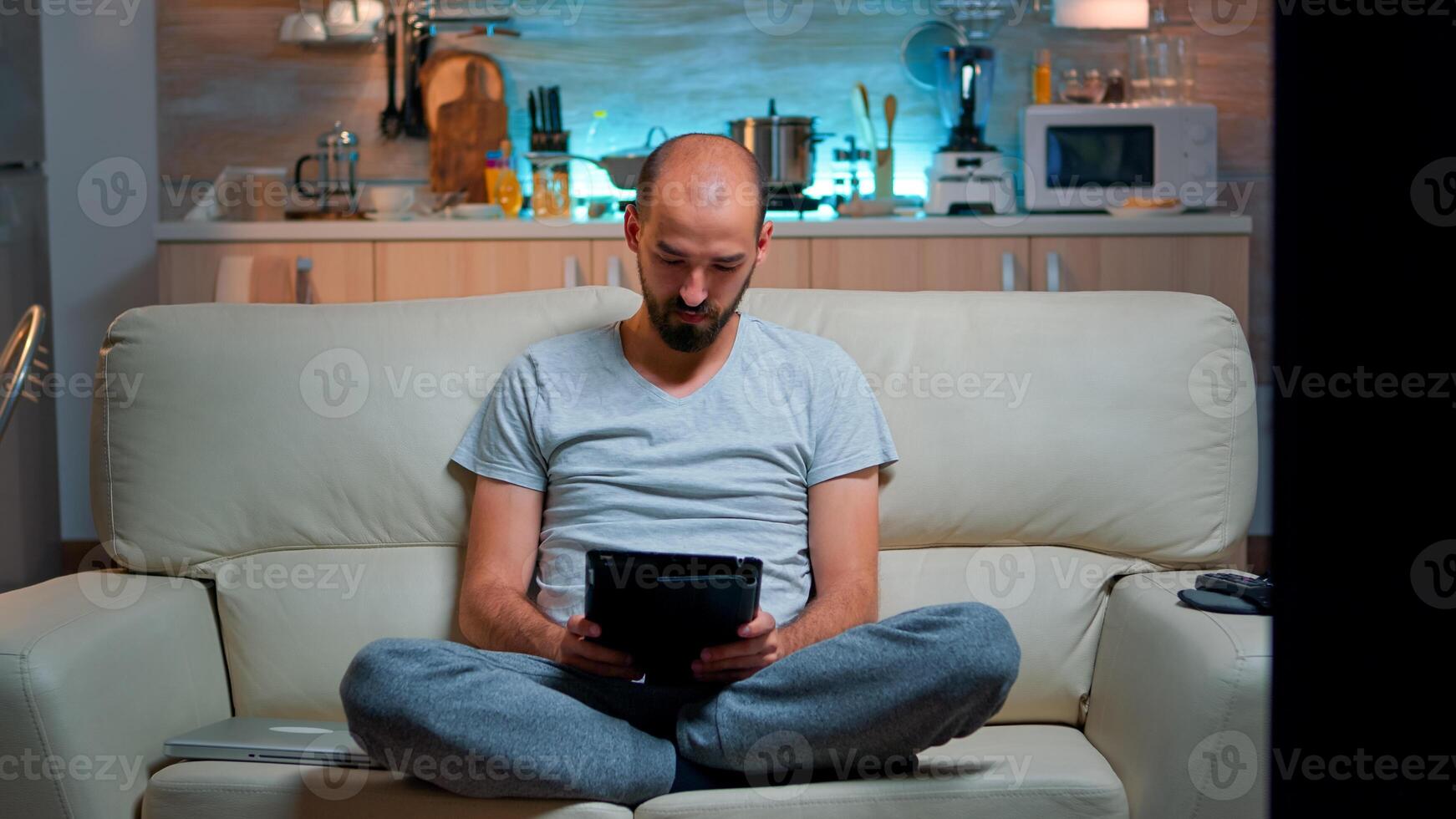 Tired man sitting alone on couch while browsing on internet using tablet computer with modern technology wireless. Caucasian male in pajamas relaxing in front of televion late at night in kitchen photo
