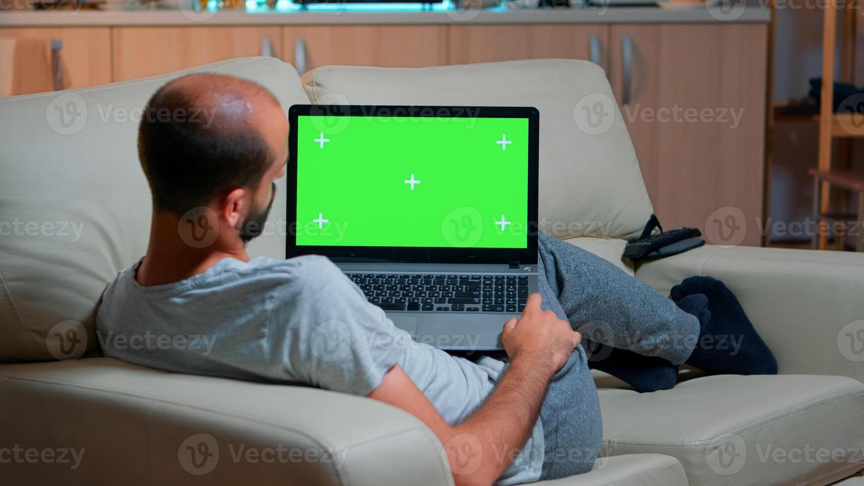 Tired man lying on sofa working on computer with mock up green screen chroma key display searching online information. Caucasian male using isolated pc late at night in kitchen photo