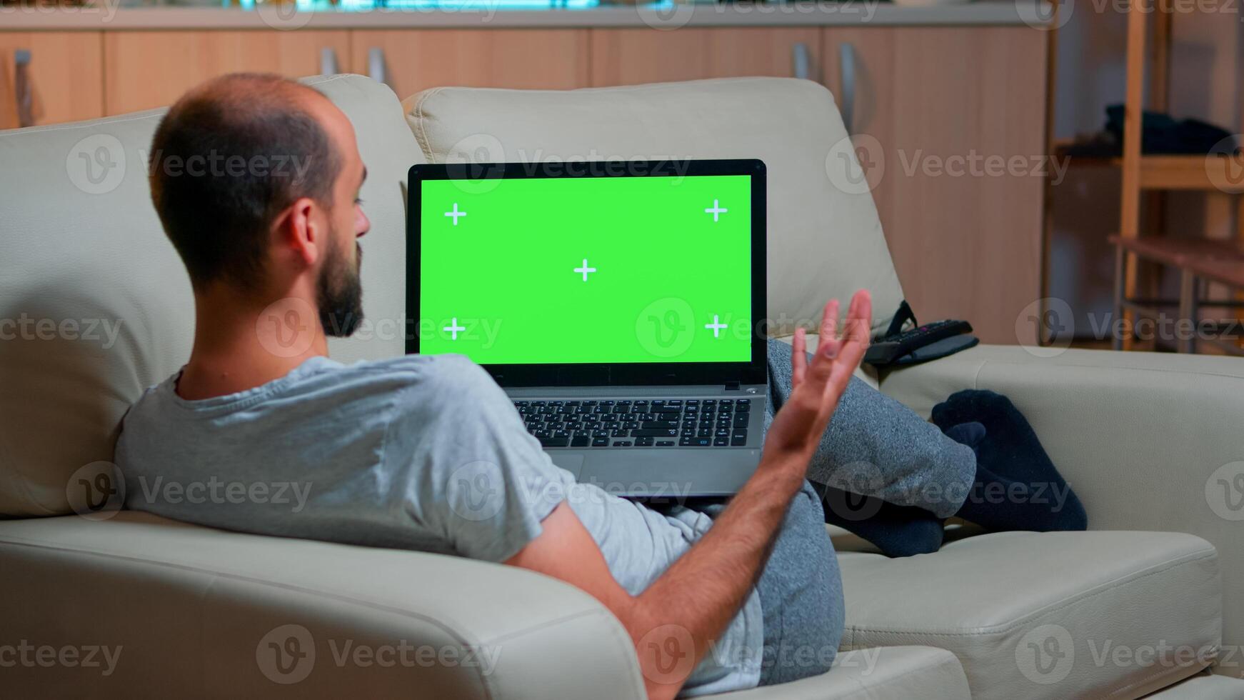 Focused man looking laptop computer with at mock up green screen chroma key display while laying on couch working at online e-learning project. Tired person using isolated pc late at night in kitchen photo