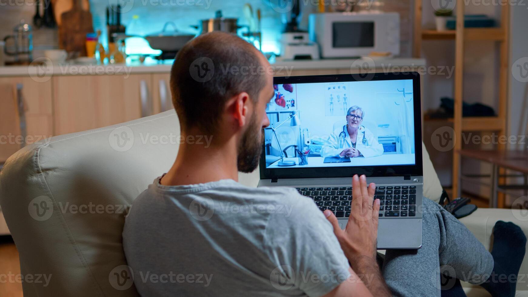 Physician doctor consulting online patient during coronavirus quarantine late at night in kitchen. Caucasian male sitting in front of laptop disscusing about illness symptoms and healthcare treatment photo
