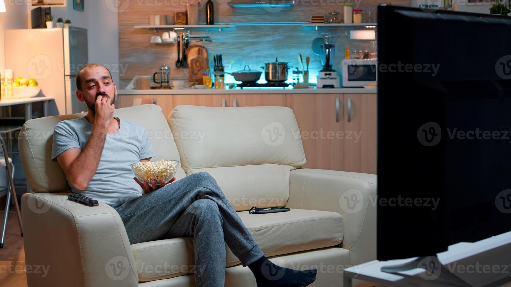 Man eating popcorn and watching TV, enjoying mid-night snack. Relaxed man lying on sofa late at night while watching entertainment tv shows at home cinema eating popcorn photo