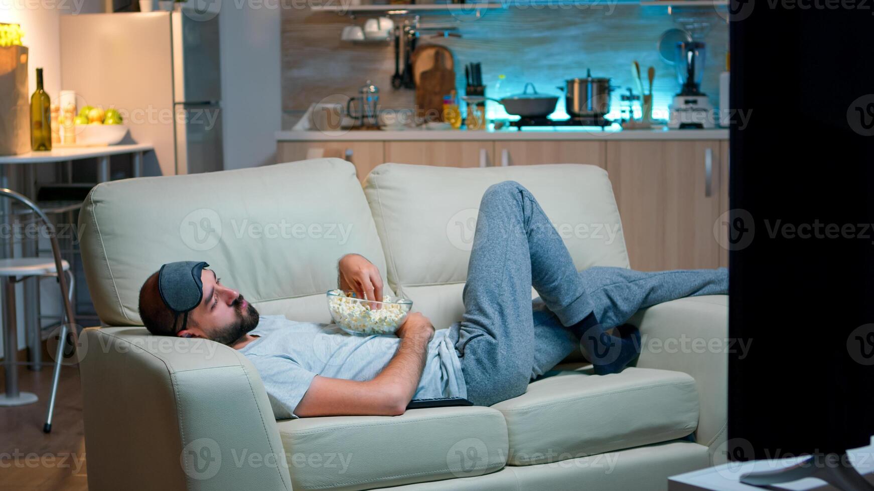 Man lying on sofa, falling asleep in fron of the TV with popcorn bowl in his hands. Tired alone man in living room after a hard day at work photo