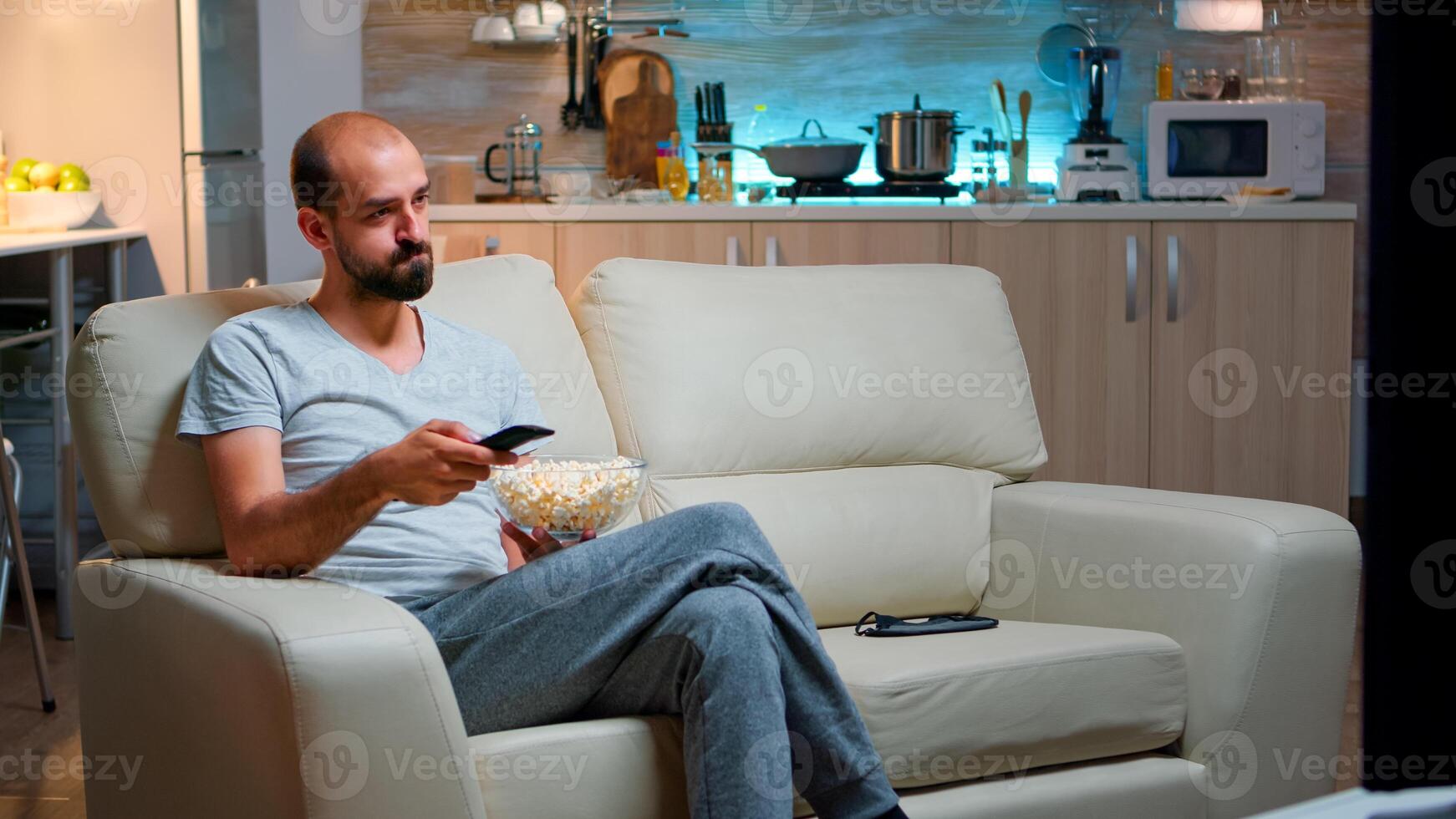 Bored man switching TV channels, holding popcorn in hads late at night while trying to watch entertainment television. Using remote control to choose favourite station and enjoy quality time photo