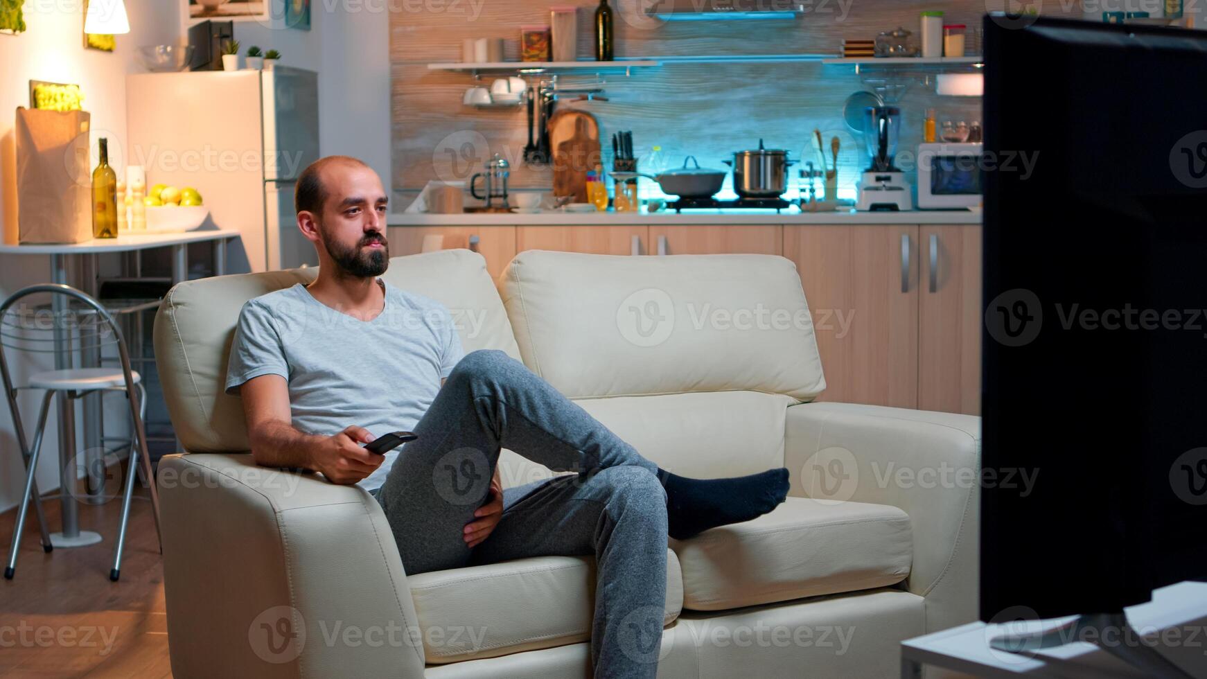 Concentrated man with beard watching movie show while lying on couch in front of television. Caucasian male in pajamas looking at entertainment shows late at night in kitchen photo