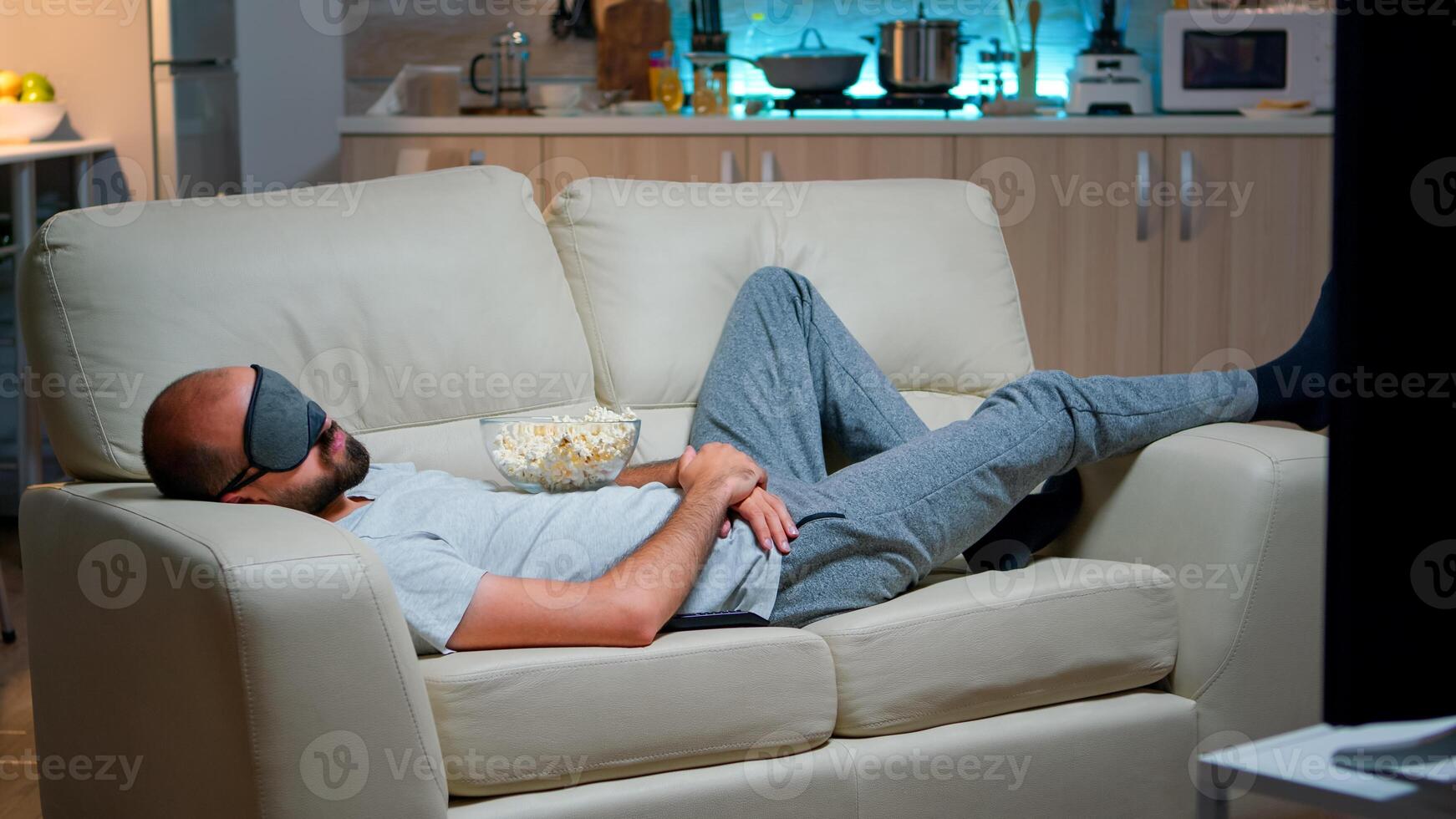 Man eye sleep mask falling asleep while watching movie, sitting in front of television. Caucasian male with beard looking entertainment series late at night in kitchen photo