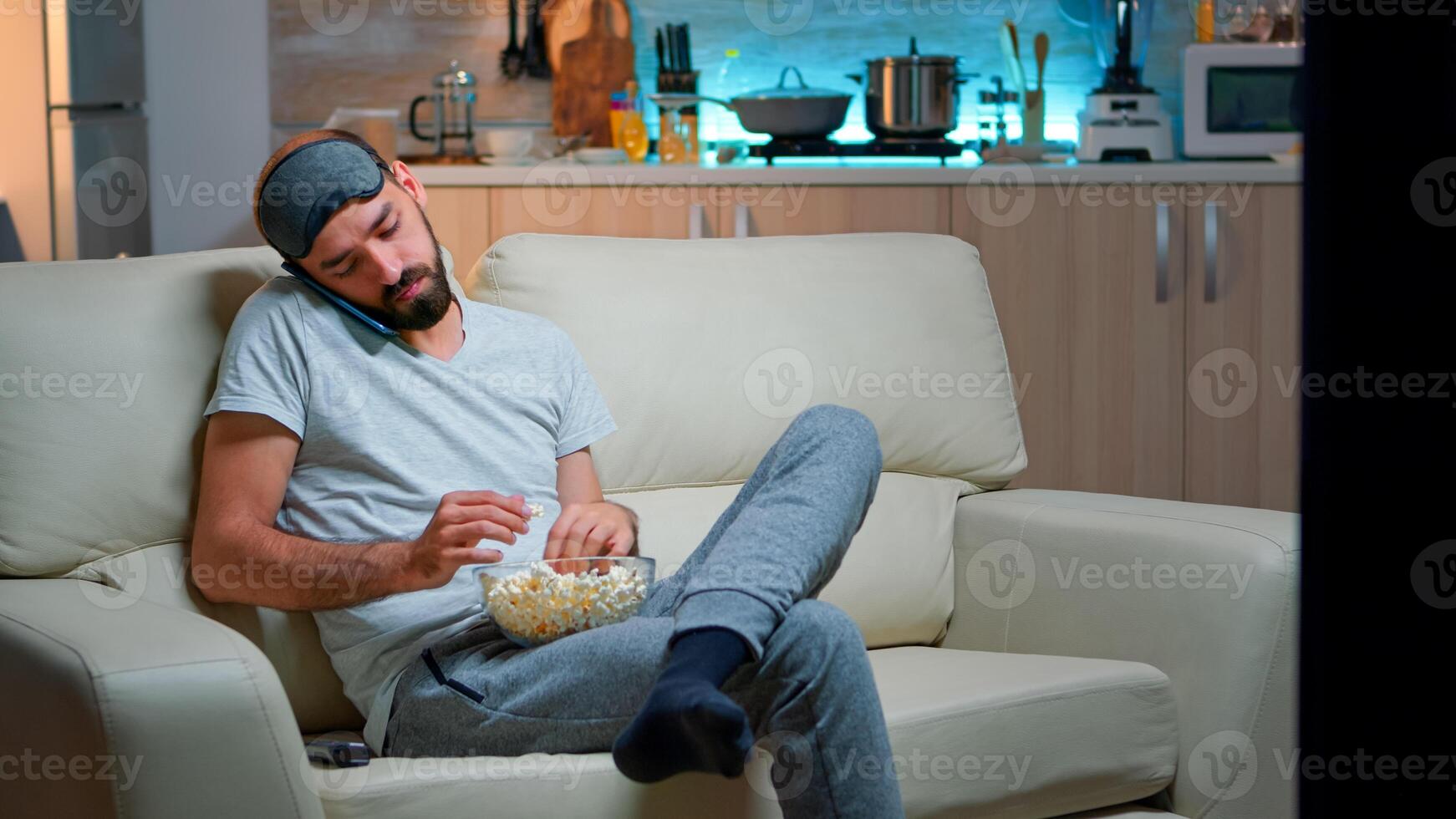 Relaxed man talking about internet connection on smartphone. Caucasian male sitting on couch watching entertainment movies while eating popcorn late at night in kitchen photo