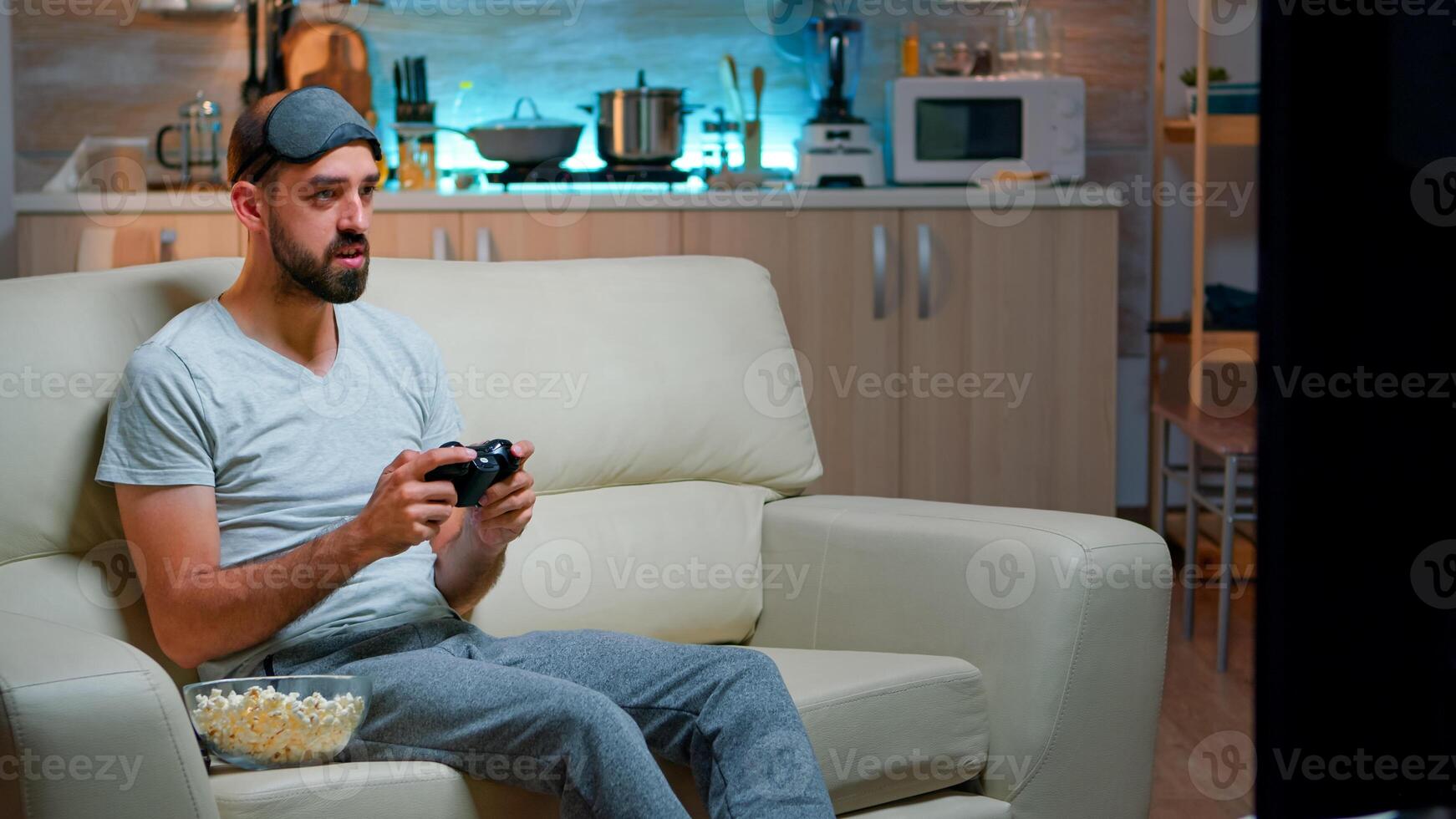 Frustrated pro gamer losing entertainment gaming competition using joystick. Serious man sitting on couch late at night in kitchen while playing online games on television photo