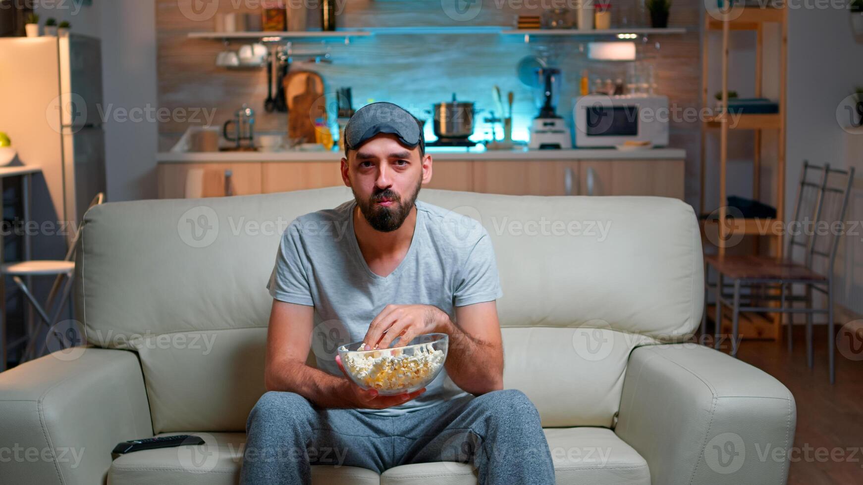 Portrait of man with beard holding popcorn bowl while watching entertainment movie on television. Concentrated person with eye sleep mask sitting on sofa late at night in kitchen photo