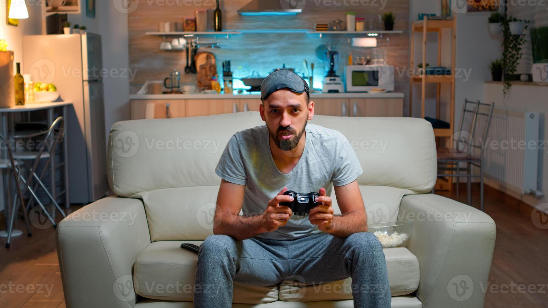 Focused man holding joystick while sitting in front of television winning online games competition. Caucasian male with sleep mask lying on couch making winner gesture late at night in kitchen photo