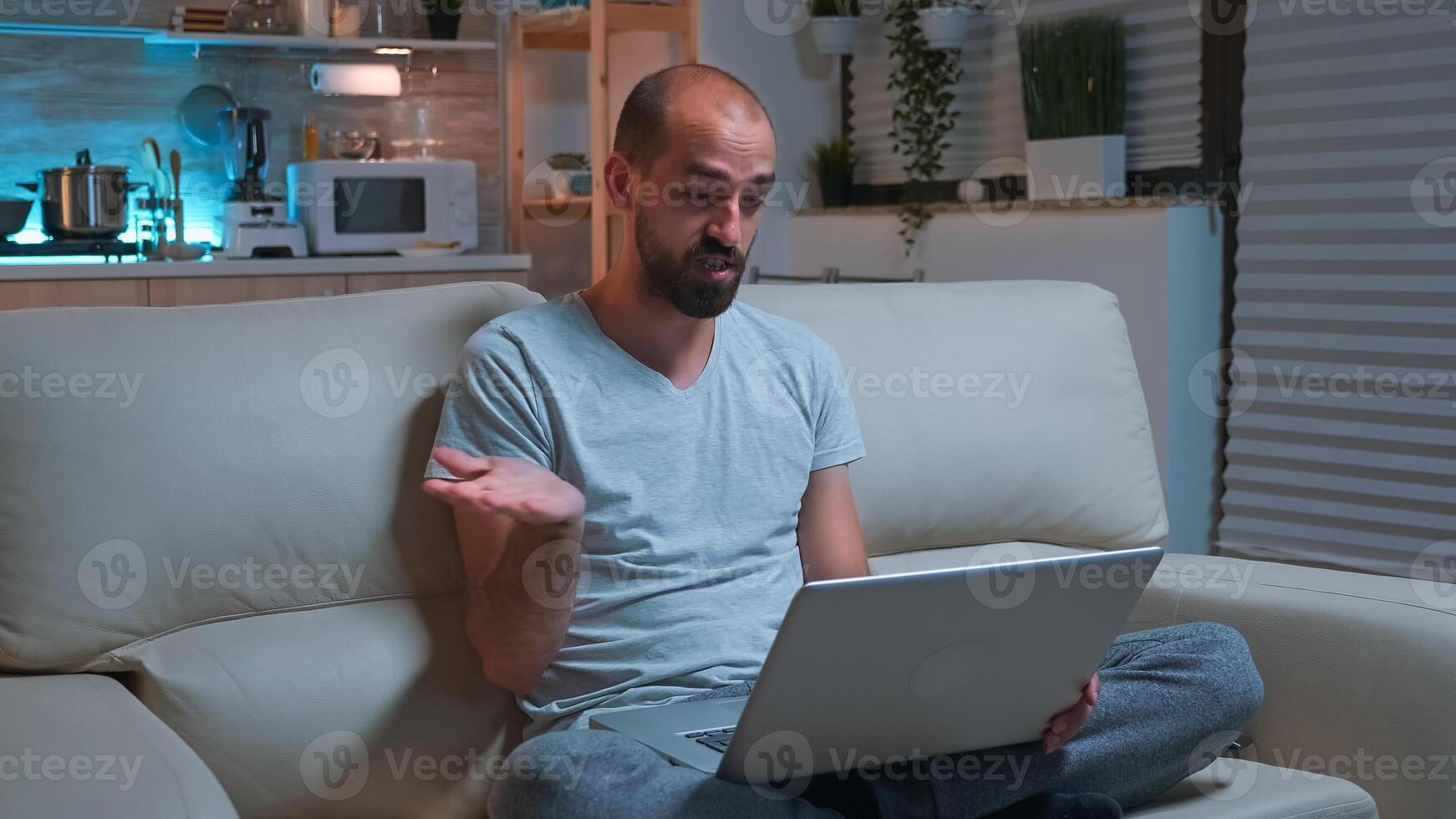 Focused man sitting on sofa in pajamas while talking with collegue during videocall conference using laptop computer. Caucasian male working at communication project late at night in kitchen photo