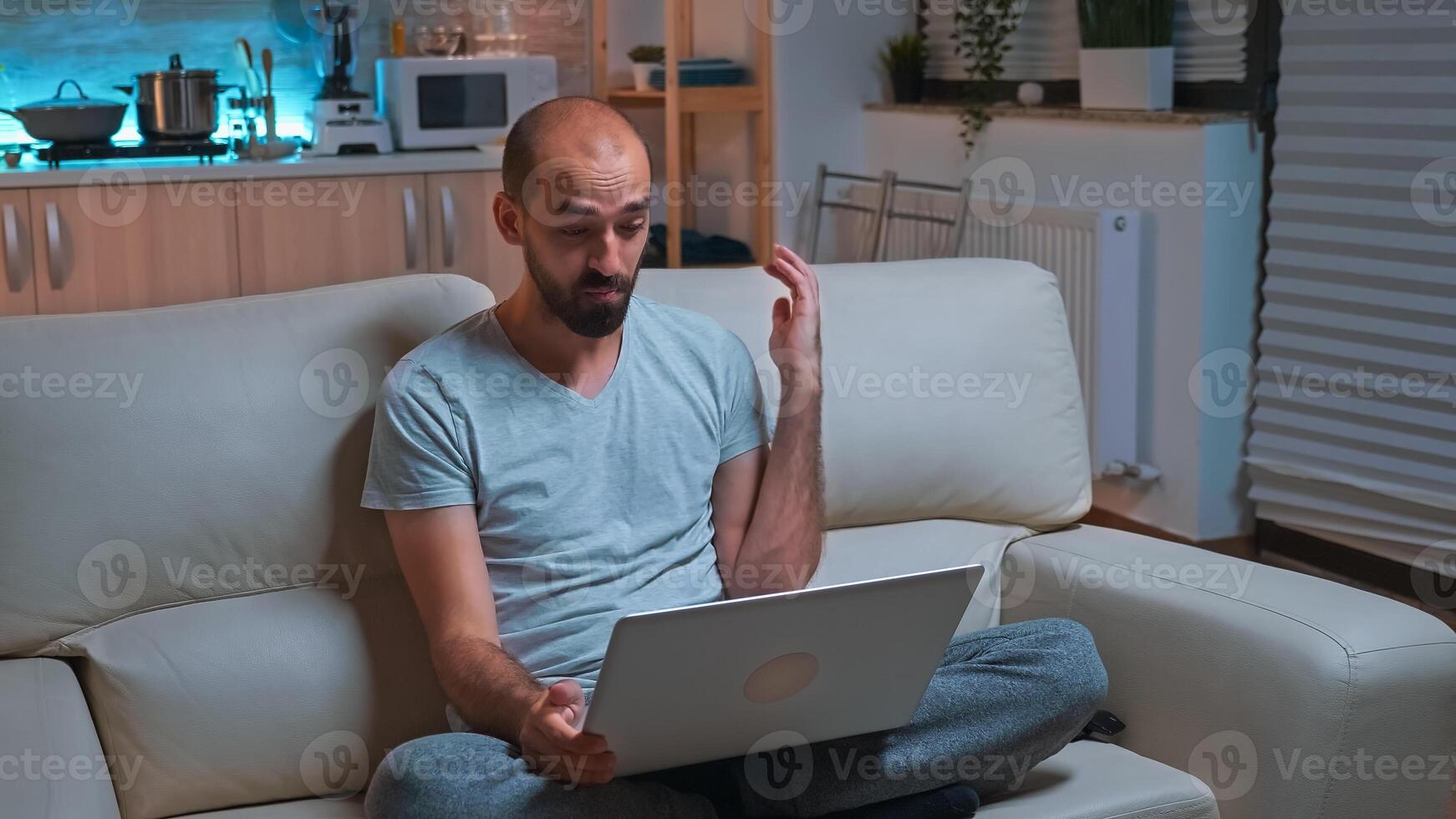 Focused man working on laptop computer during business videocall conference. Caucaisan male sitting on sofa in pajamas late at night in kitchen using modern technology wireless photo