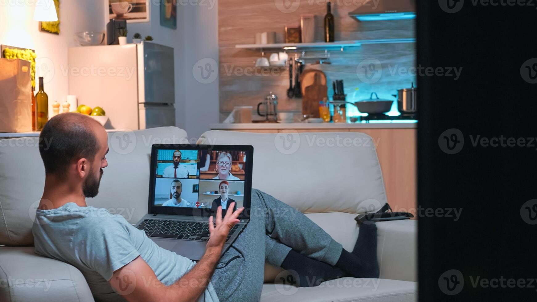 Tired man relaxing on sofa in pajamas during online vidoecall meeting using laptop computer. Caucasian male working at social media project from home because of coronavirus quarantine late at night photo