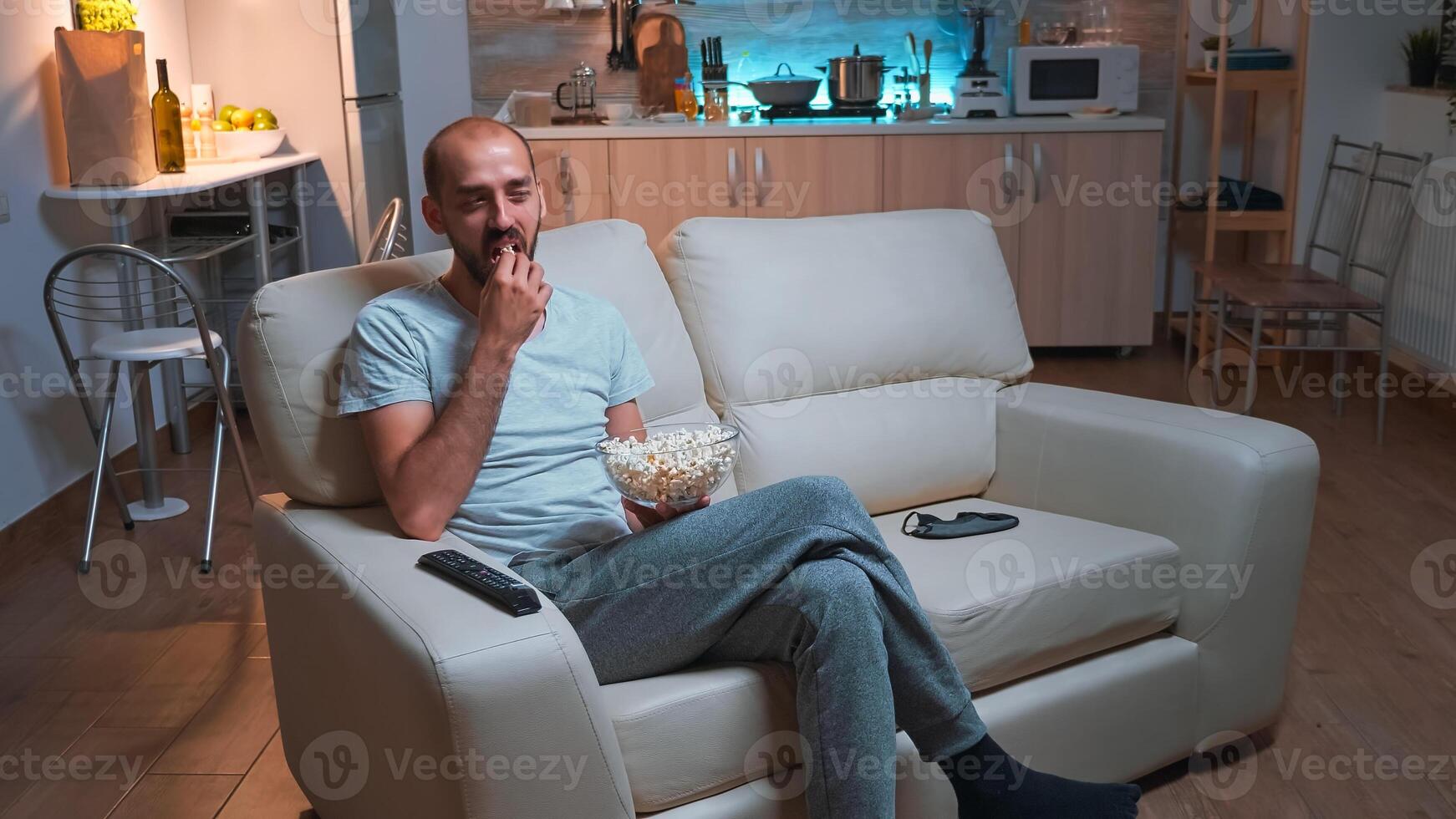 Man with beard watching tv show using remote control to change channels on television. Caucasian male relaxing on couch with popcorn bowl in hands looking entertainment series at late at night photo