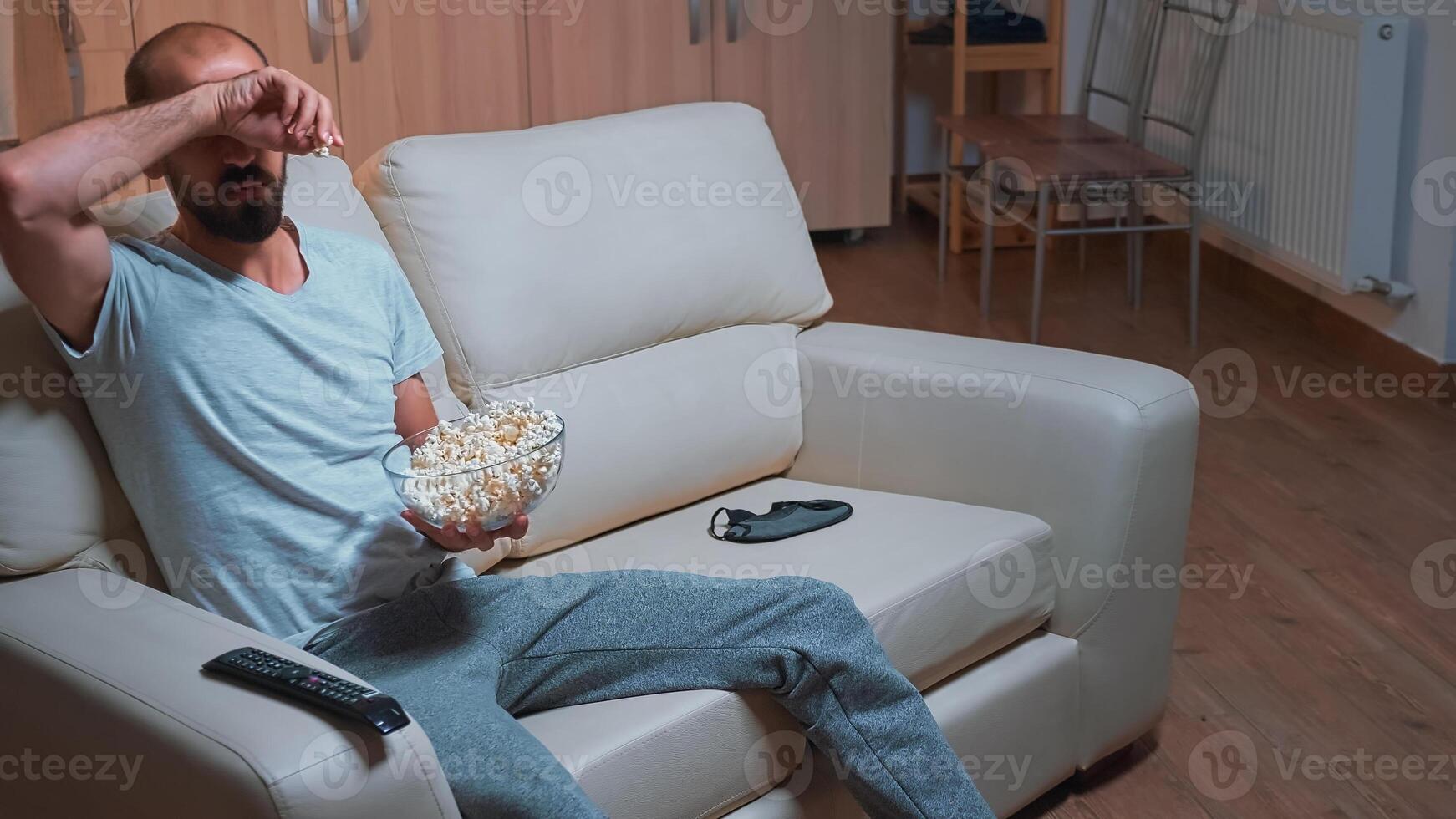 Concentrated man chilling on sofa in front of television watching movie series while eating popcorn. Caucasian male in pajamas looking at entertainment shows late at night in kitchen photo