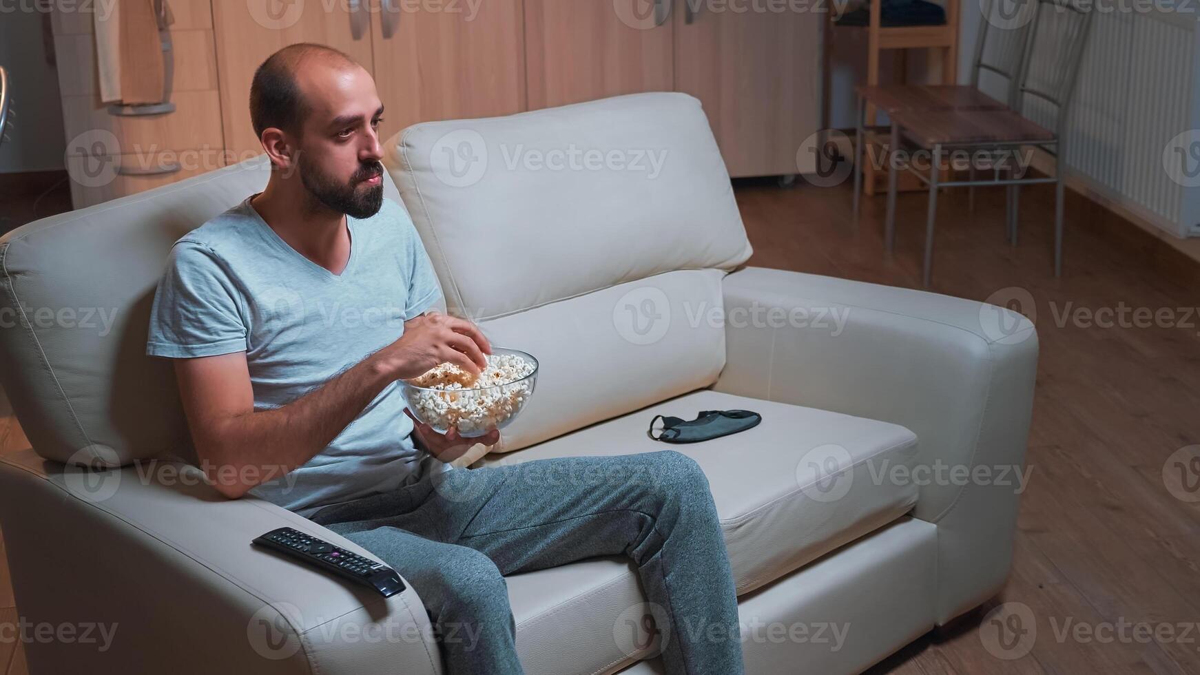 Caucasian male sitting on couch with popcorn bowl in hands while looking at movie series on television. Tired man in pajamas watching at entertainment shows late at night in kitchen photo