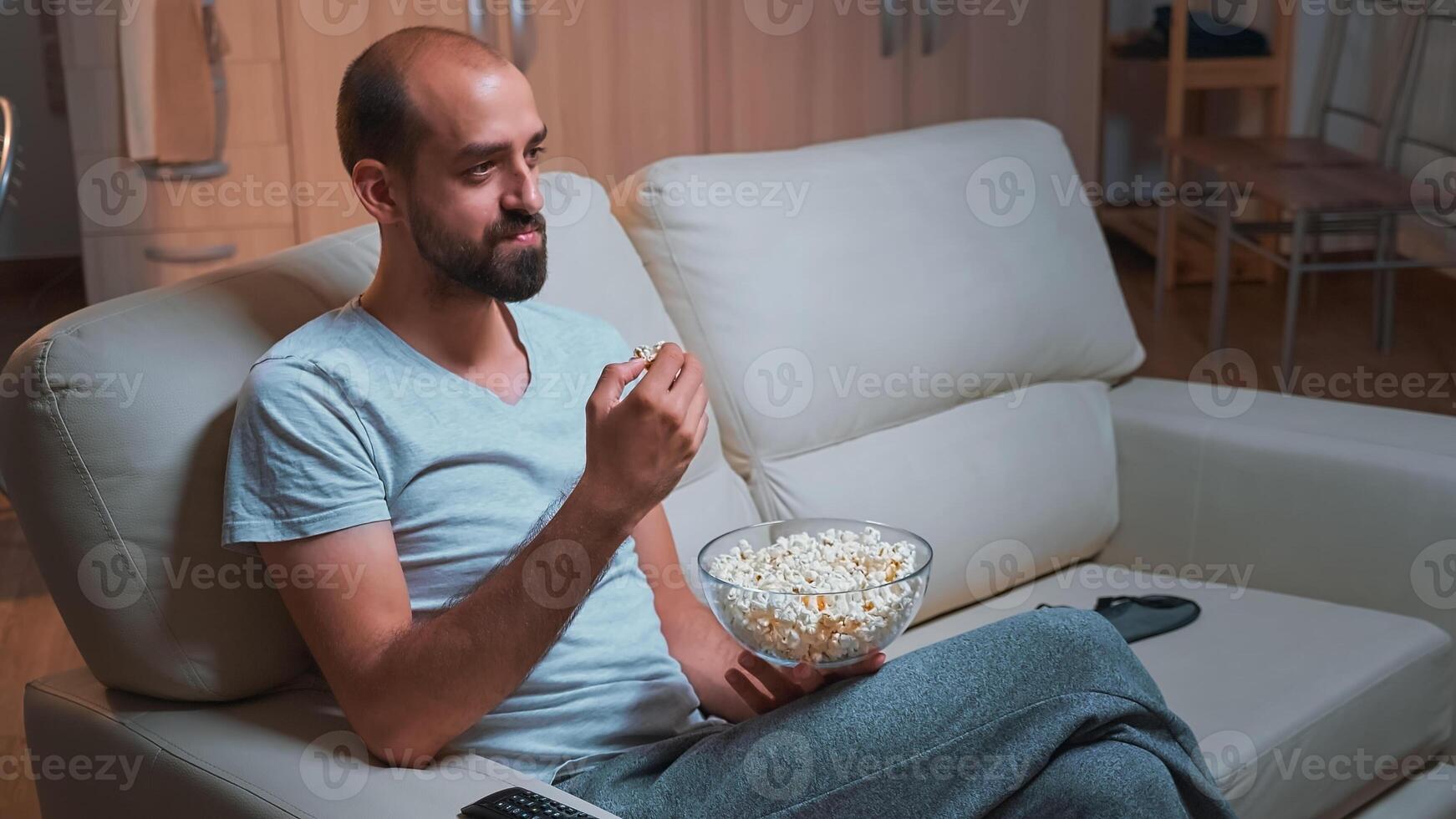 Caucasian male chilling on sofa with popcorn bowl in hands while watching movie series on television. Tired man in pajamas looking at entertainment shows late at night in kitchen photo