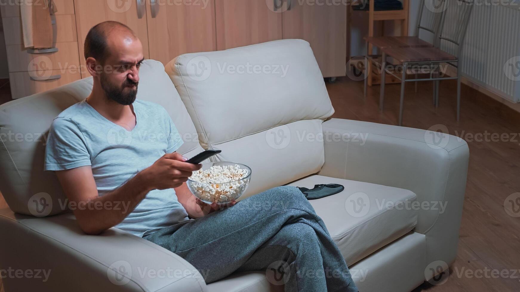 Concentrated man sitting in front of television using control remote while watching movie film. Caucasian male in pajamas looking at entertainment shows late at night in kitchen photo