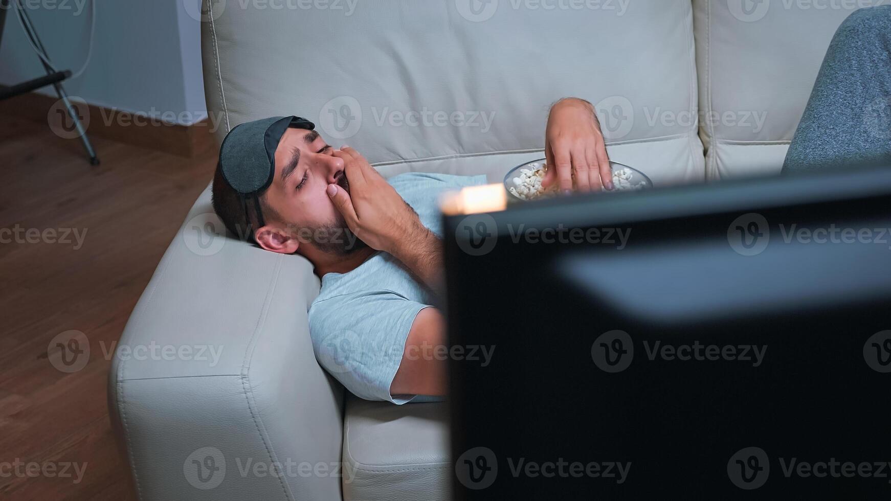 Caucasian male with eye sleep mask eating popcorn while watching entertainment tv show. Tired man sitting in front of television during movie film late at night in kitchen photo
