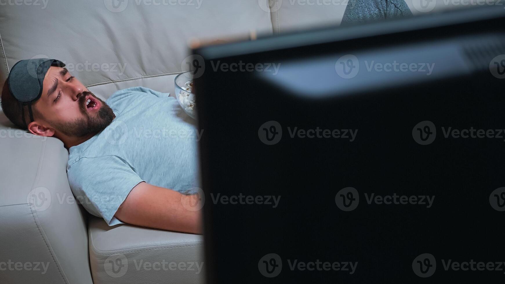Man with eye sleep mask lying on sofa while watching entertainment movie holding popcorn bowl in hands. Caucasian male sitting in front of television during tv show late at night in kitchen photo