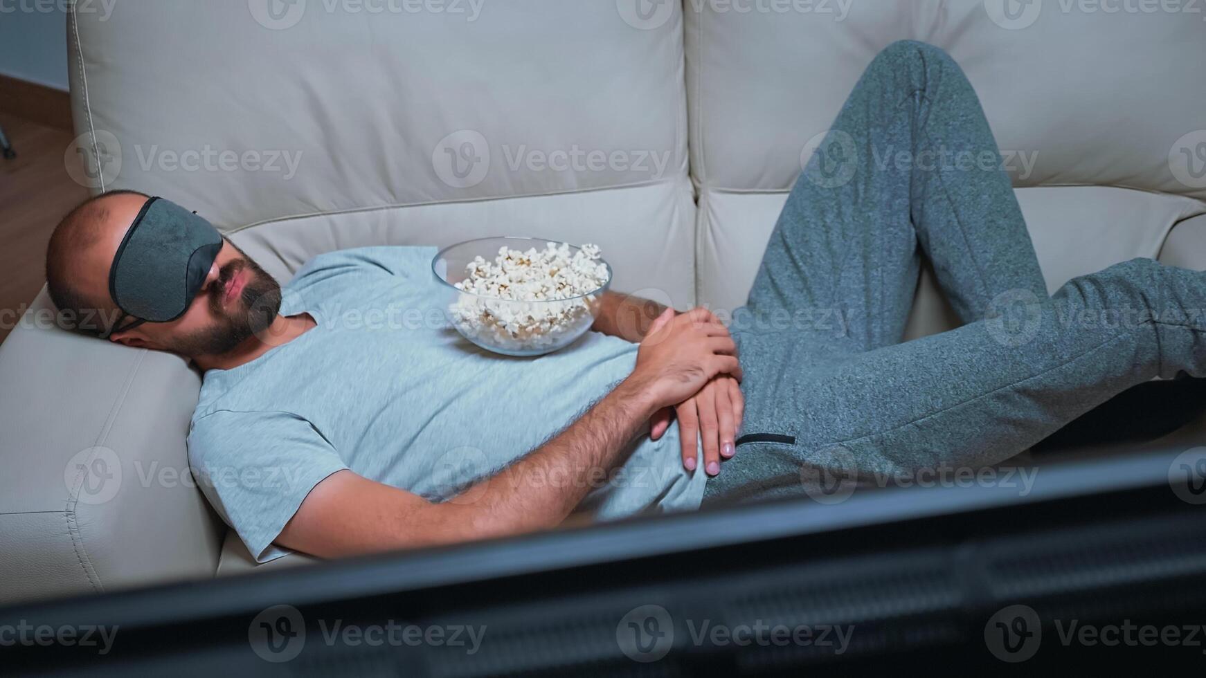 Caucasian male falling asleep while watching movie show sitting on couch late at night in kitchen. Tired man wearing eye sleep mask while the tv is on photo