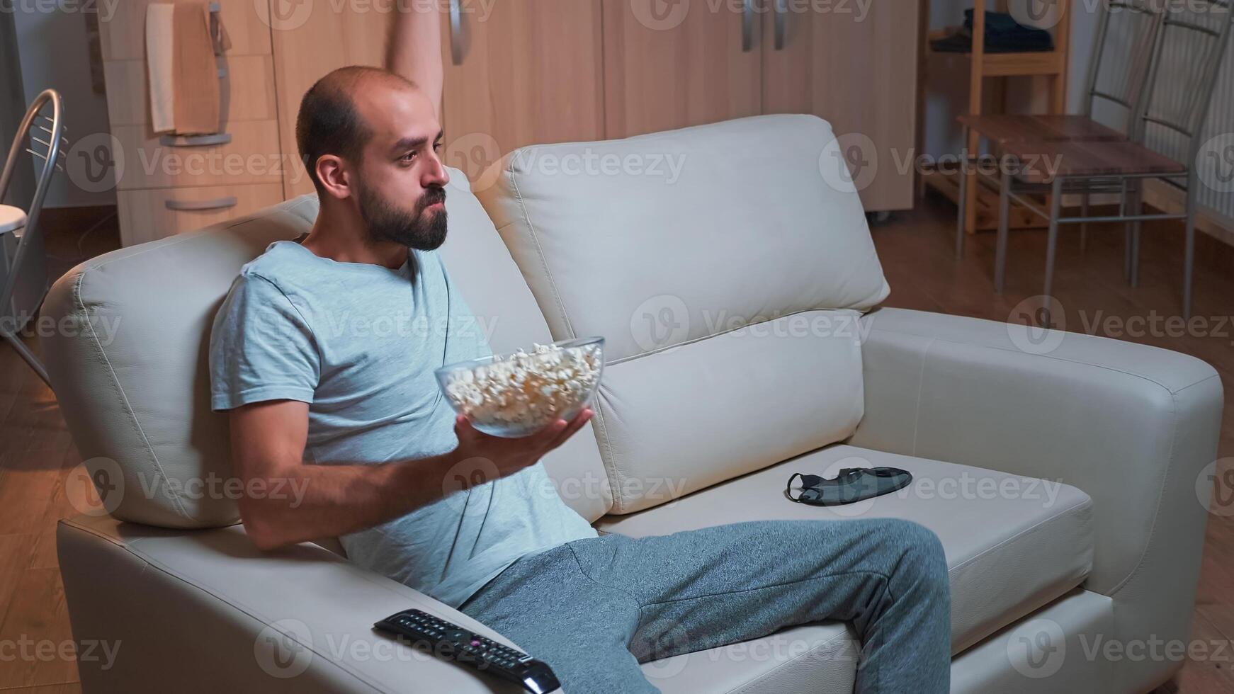 Concentrated man watching entertainment movie while making face expression, holding popcorn bowl in hands. Freelancer in pajamas sitting on sofa looking at entertainment shows late at night in kitchen photo