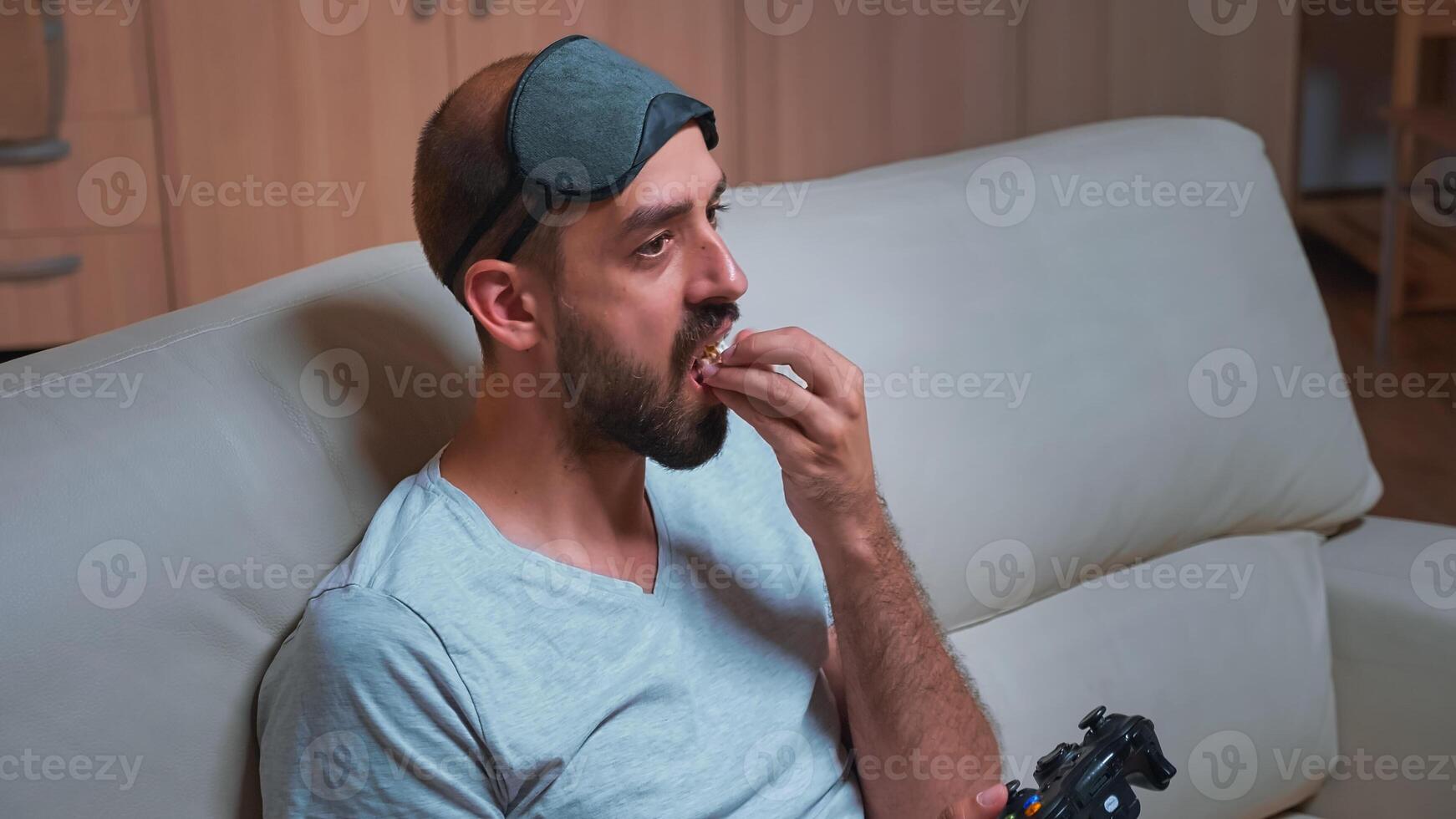 Pro gamer with sleep mask sitting in front of television playing videogames using joystick during quarantine. Player man enjoying free time on couch eating popcorn late at night in kitchen photo
