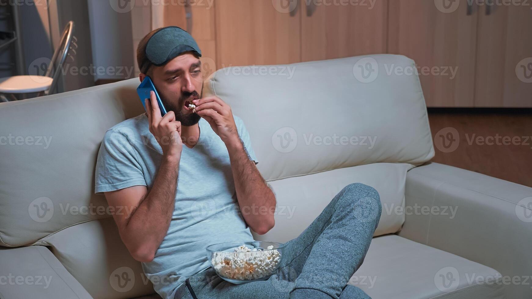 Caucasian male resting on couch eating popcorn while talking on phone with friend about social network. Relasex man sitting in front of television watching entertainment show late at night in kitchen photo