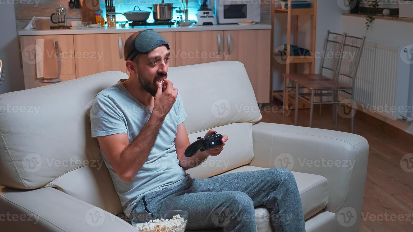 Frustrated man with sleep mask losing videogame competition using wireless controller. Disappointed pro gamer lying on sofa in front of television late at night in kitchen photo