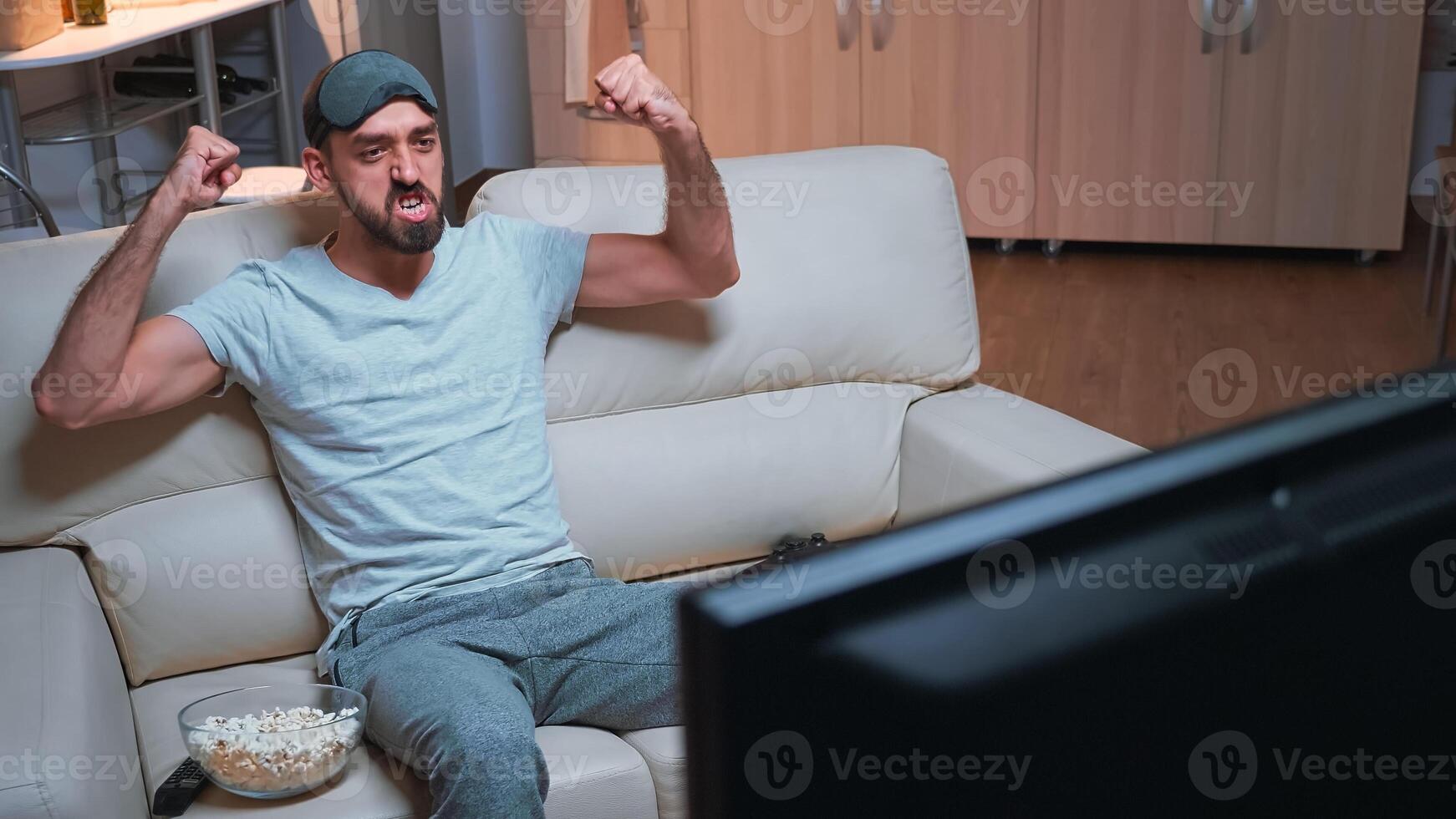 Player man holding wireless controller while playing videogames during quarantine. Pro gamer sitting in front of television on sofa making happy gesture late at night in kitchen photo