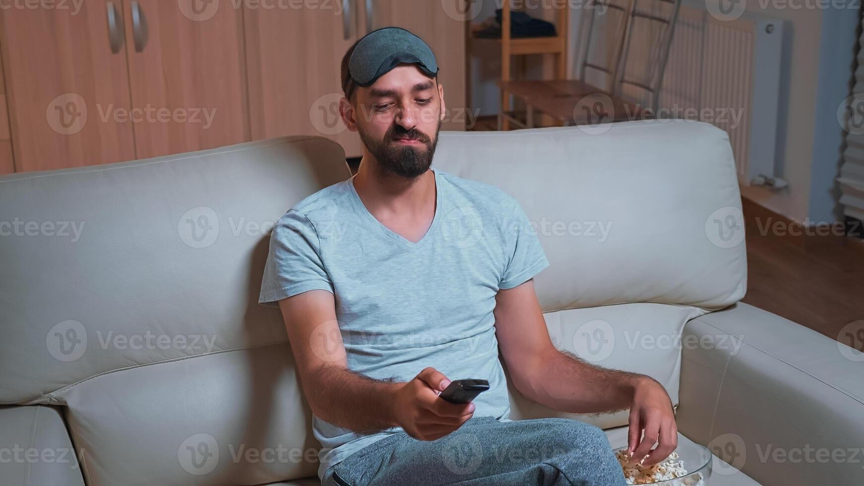 Interested man with beard relaxing in front of television while looking at entertainment movie using remote controle. Caucasian male lying on sofa eating popcorn late at night in kitchen photo