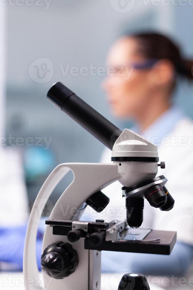 Selective focus on medical microscope standing on table in pharmacology microbiologist hospital laboratory. Specialist doctor typing disease expertise on computer discovering genetic substance photo
