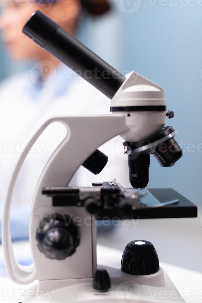 Selective focus on medical microscope standing on table in pharmacology microbiologist hospital laboratory. Specialist doctor typing disease expertise on computer discovering genetic substance photo