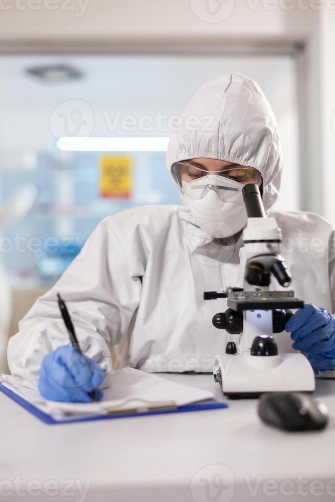 Doctor writing notes dressed in ppe suit and visor mask using microscope. Chemist in coverall working with various bacteria, tissue blood samples for antibiotics research. photo
