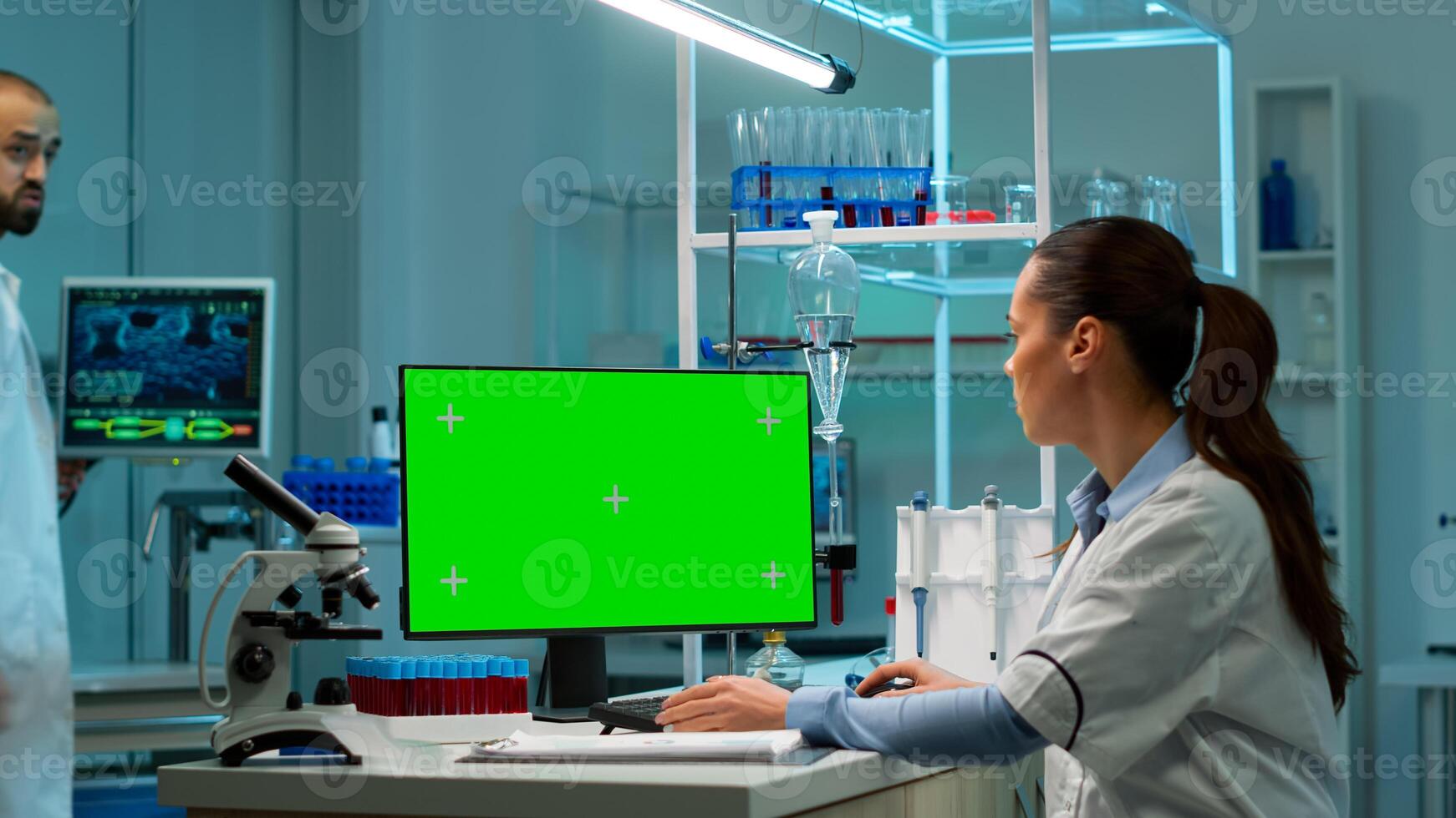 Scientist sitting at desk working on personal computer with mock-up green screen. In background man lab researcher discussing with doctor about vaccine developent bringing blood samples photo