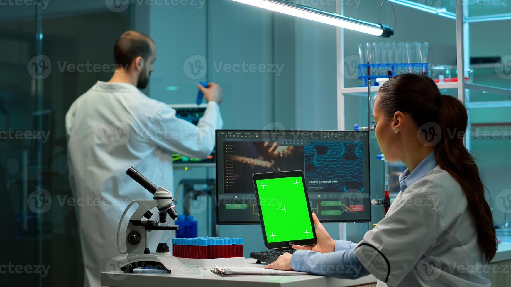 Microbiologist working on notepad with green chroma key display in modern equipped lab. Team of biotechnology scientists developing drugs using tablet with mock up screen. photo