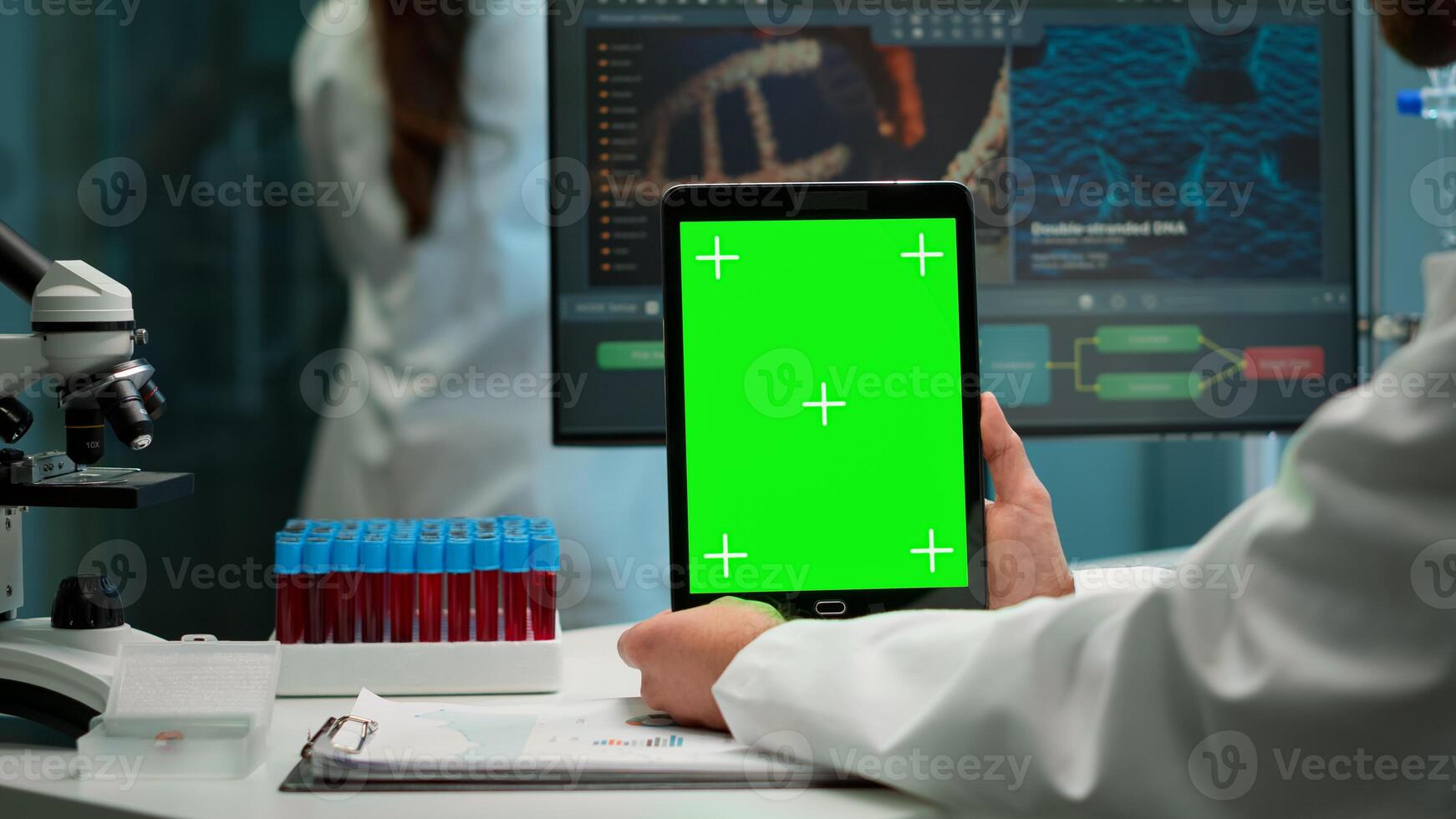 Close up of biochemist sitting at workplace in laboratory using green mock-up screen tablet with chroma key display. coworker working in background bringing blood sample. photo